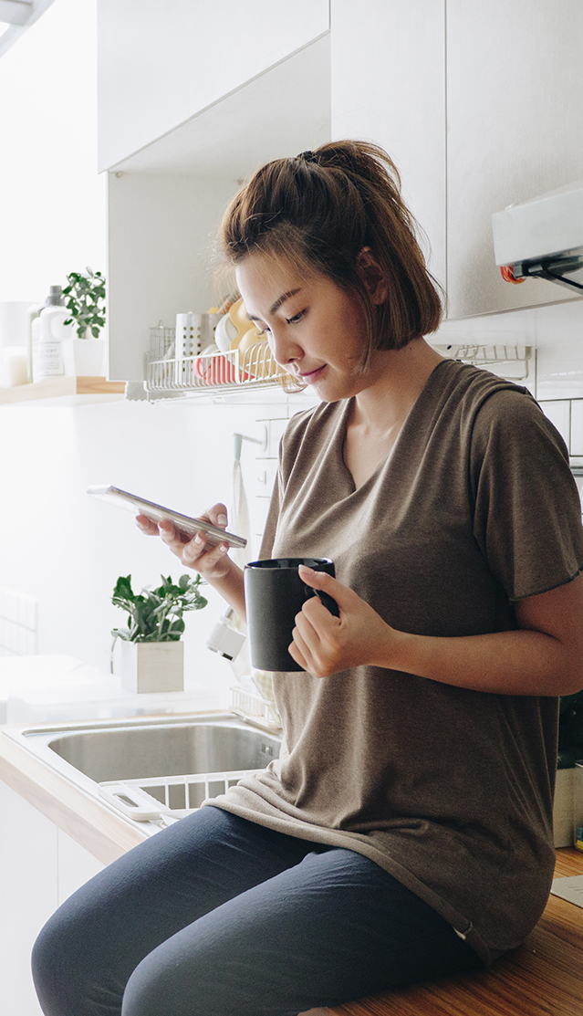 woman sitting on countertop holding mug while looking at phone